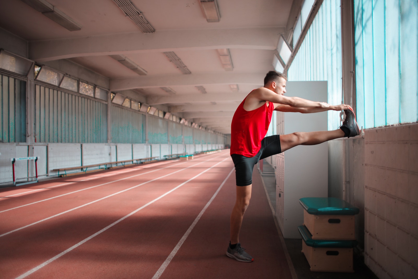 Side View Photo of Man in Red Tank Top and Black Shorts Stretching in Indoor Running Track